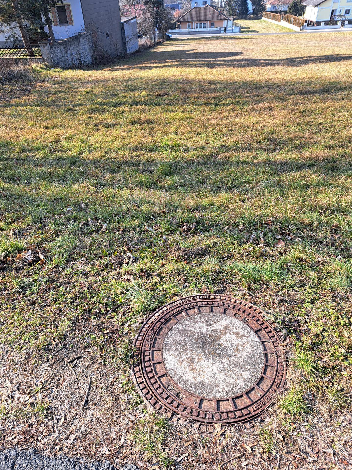 Manhuldeckel auf dem Boden eines Grundstücks mit Blick auf die umliegende Bebauung und Landschaft.