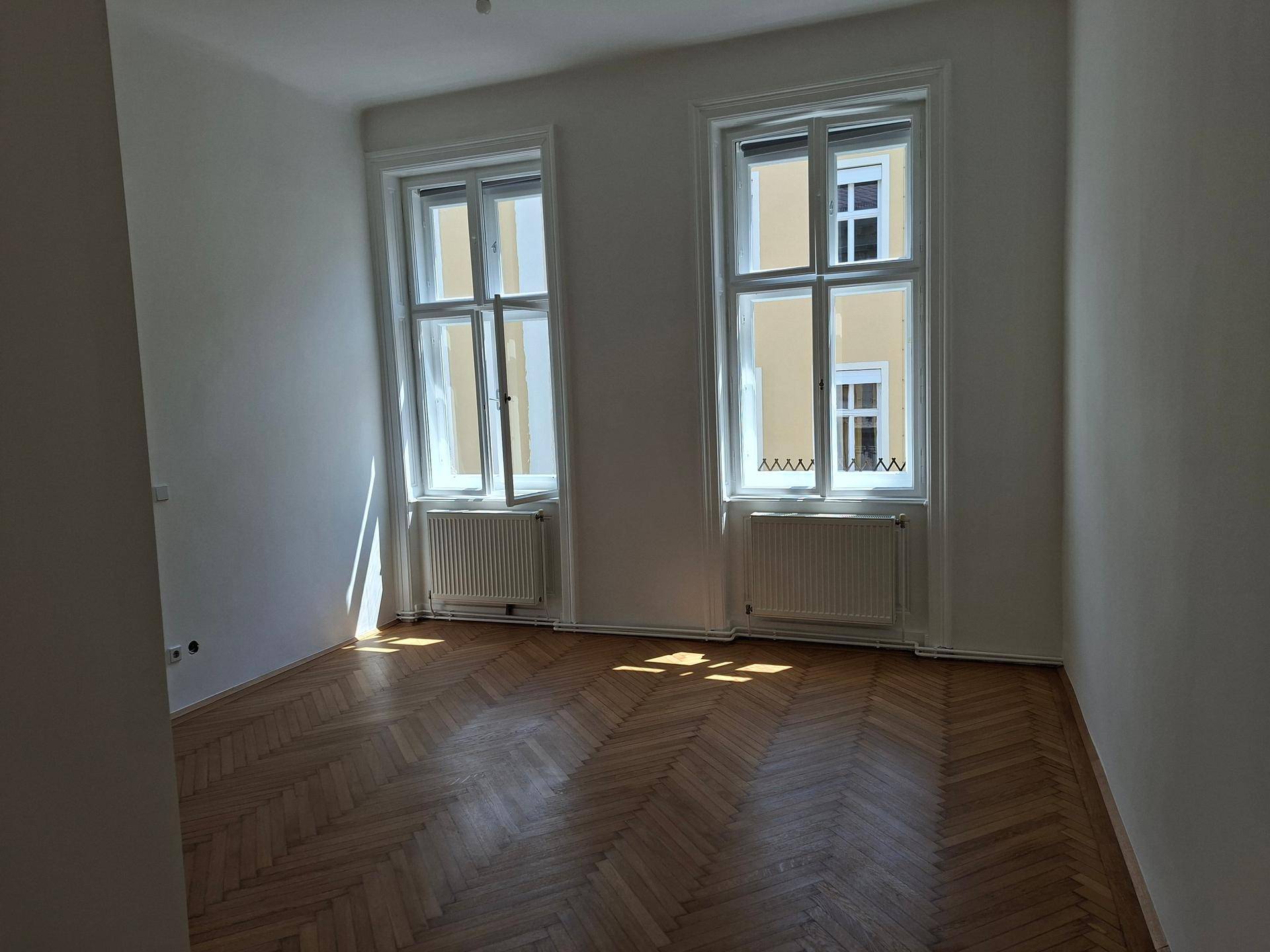 Geräumiges Zimmer mit Fischgrätparkett und zwei großen Fenstern, die den Raum erhellen.