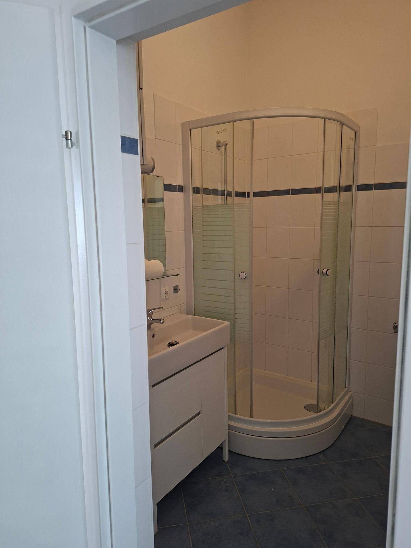 Kleines Badezimmer mit Duschkabine, Waschbecken und weißen Fliesen mit blauen Akzenten.