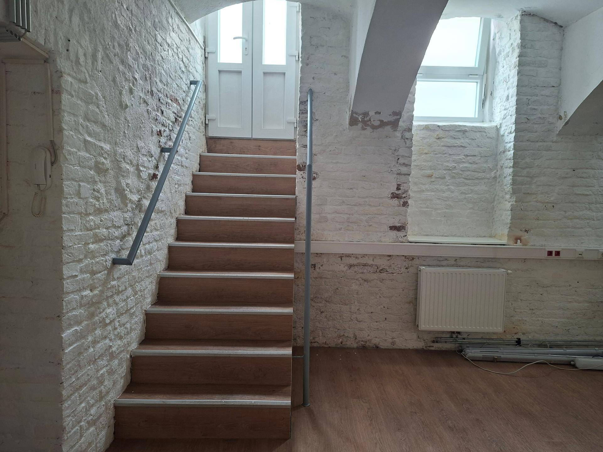 Holztreppe mit Metallgeländer, umgeben von weiß gestrichenen Ziegelwänden und Fenstern.