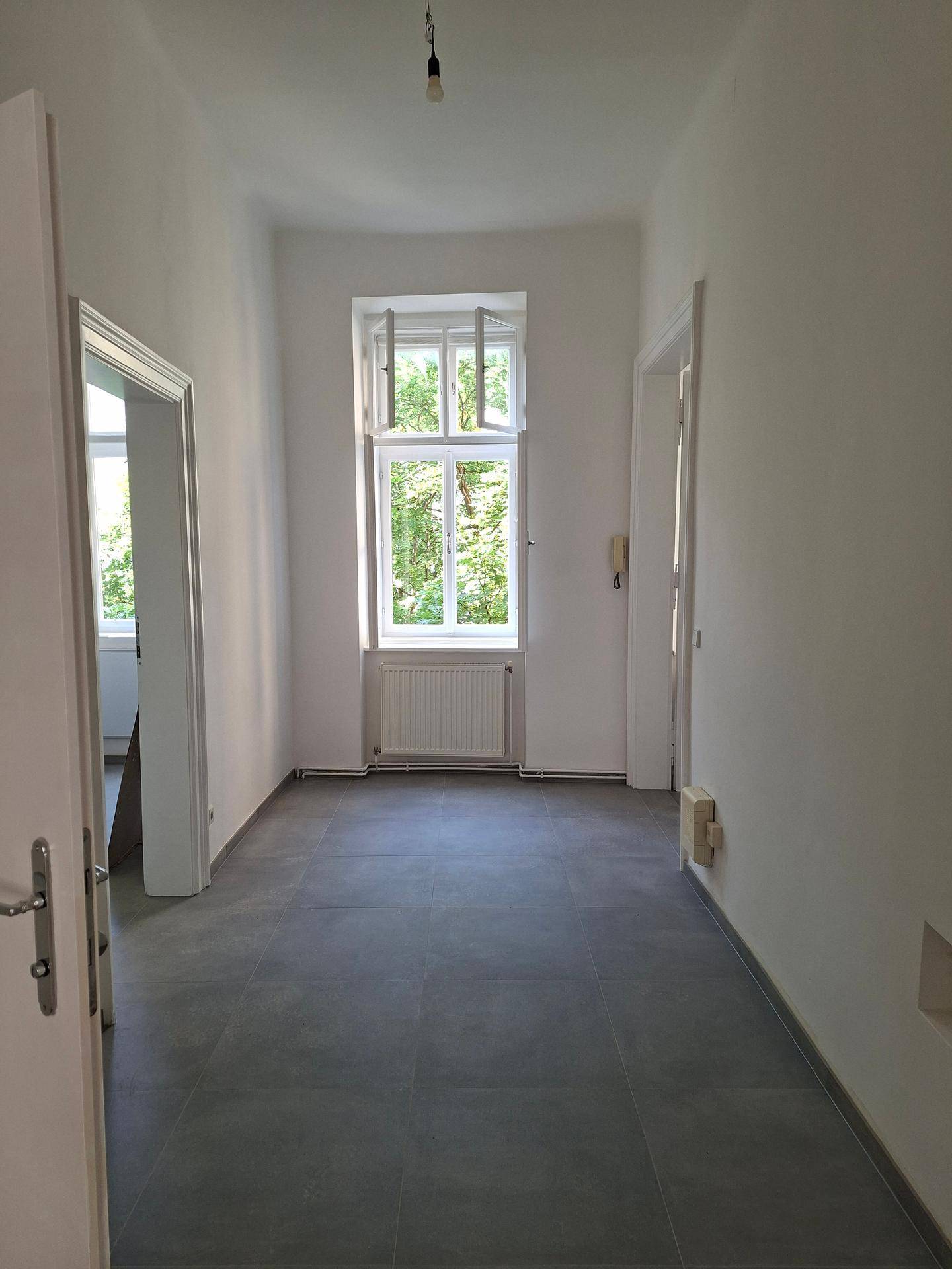 Langer, heller Flur mit grauen Fliesen und einem Fenster mit Blick ins Grüne.