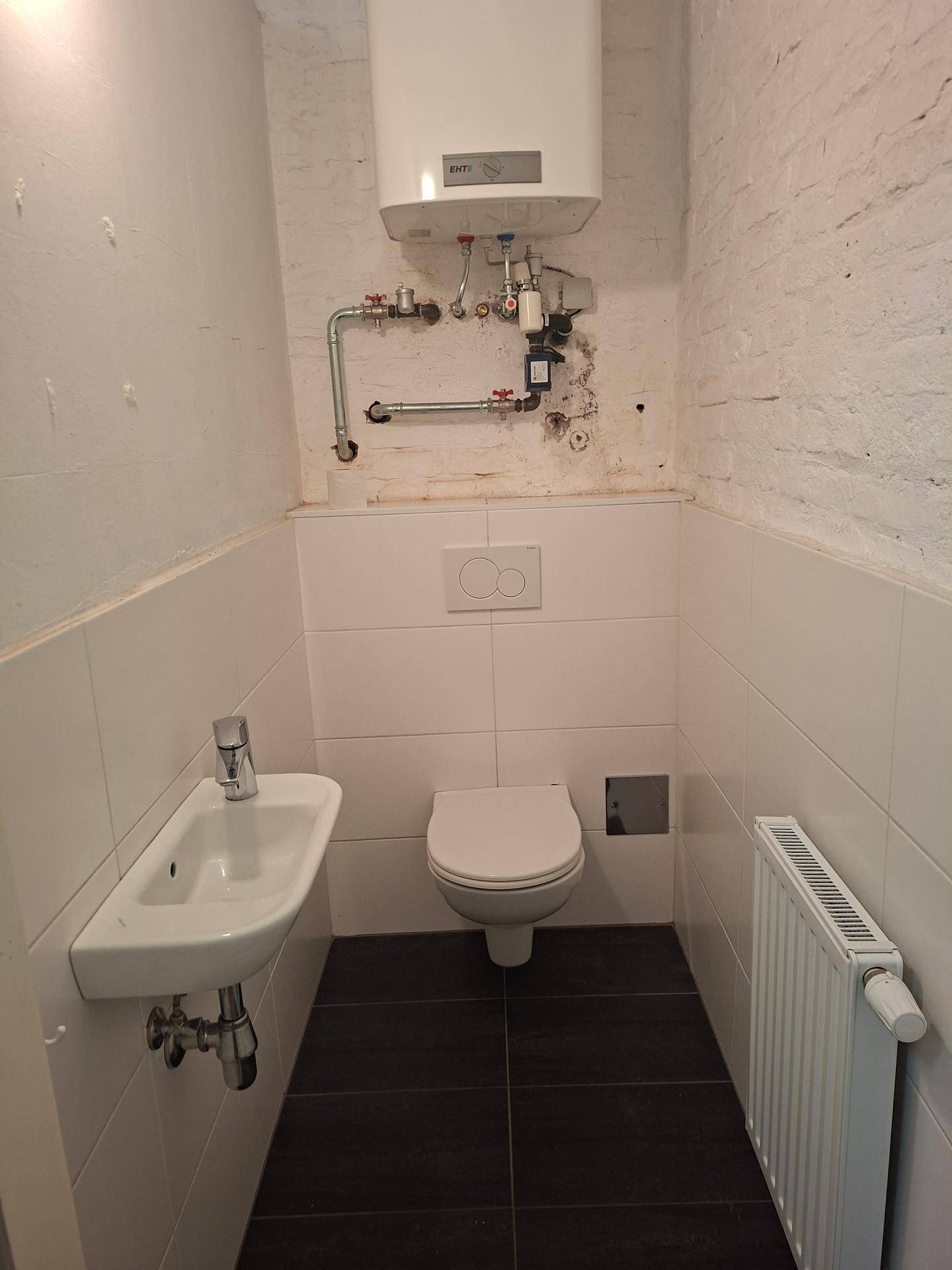 Kleine Gästetoilette mit wandhängendem WC, Waschbecken und Warmwasserbereiter an der Wand.