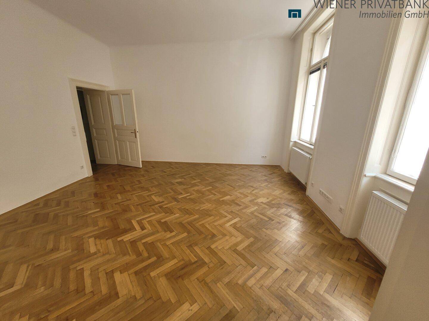 Geräumiges Zimmer mit klassischem Fischgrätparkett und hohen Fenstern.