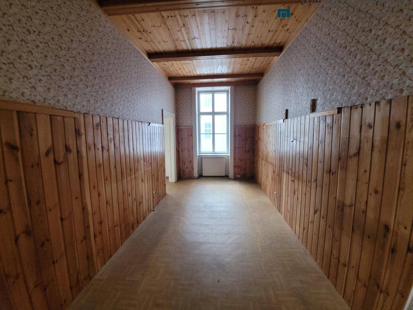Langer Raum mit Holzvertäfelung, Holzbalkendecke und Fenster, in sanierungsbedürftigem Zustand.