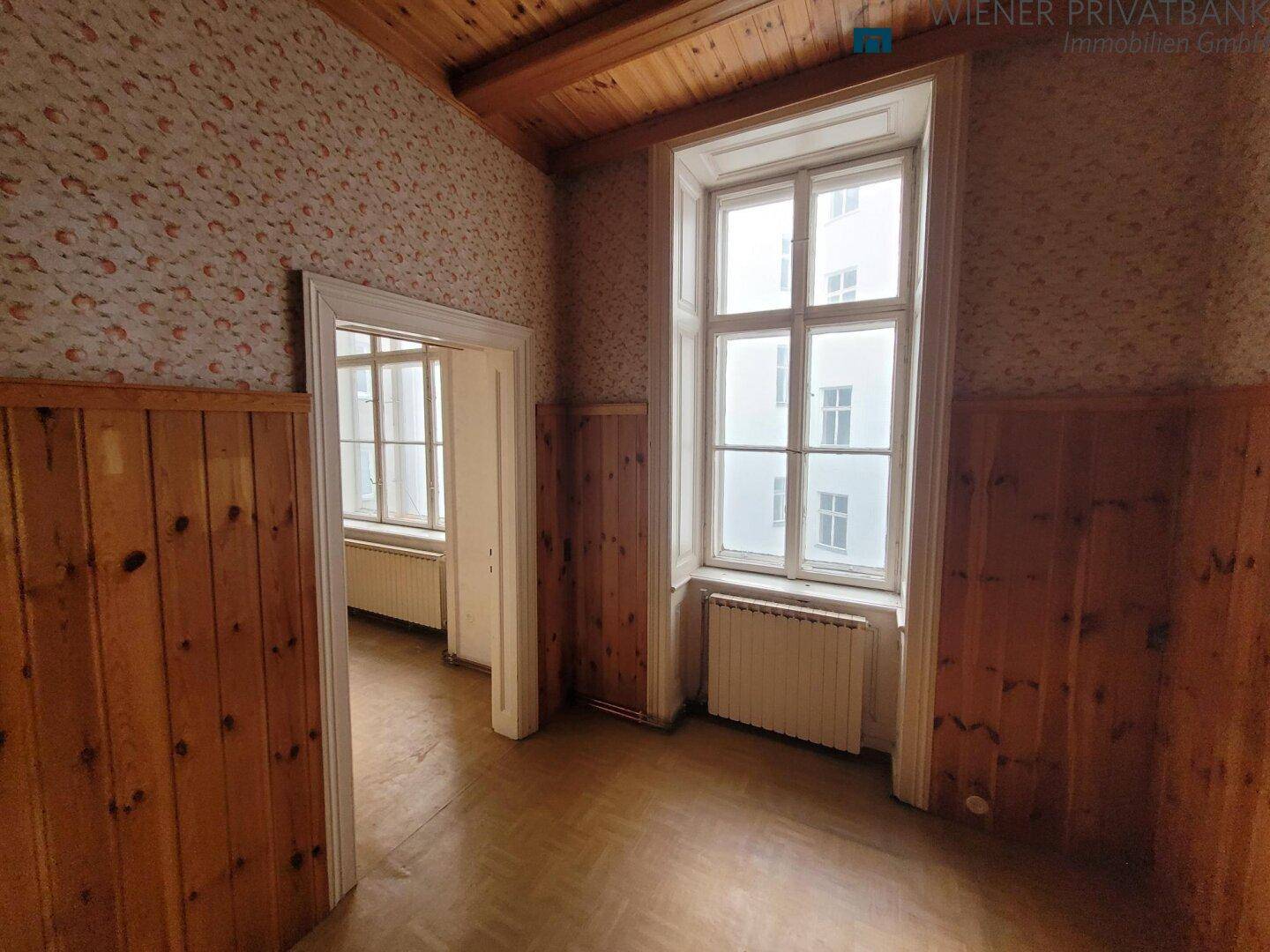 Innenraum mit rustikaler Holzvertäfelung, Holzbalkendecke und zwei großen Fenstern, renovierungsbedürftig.