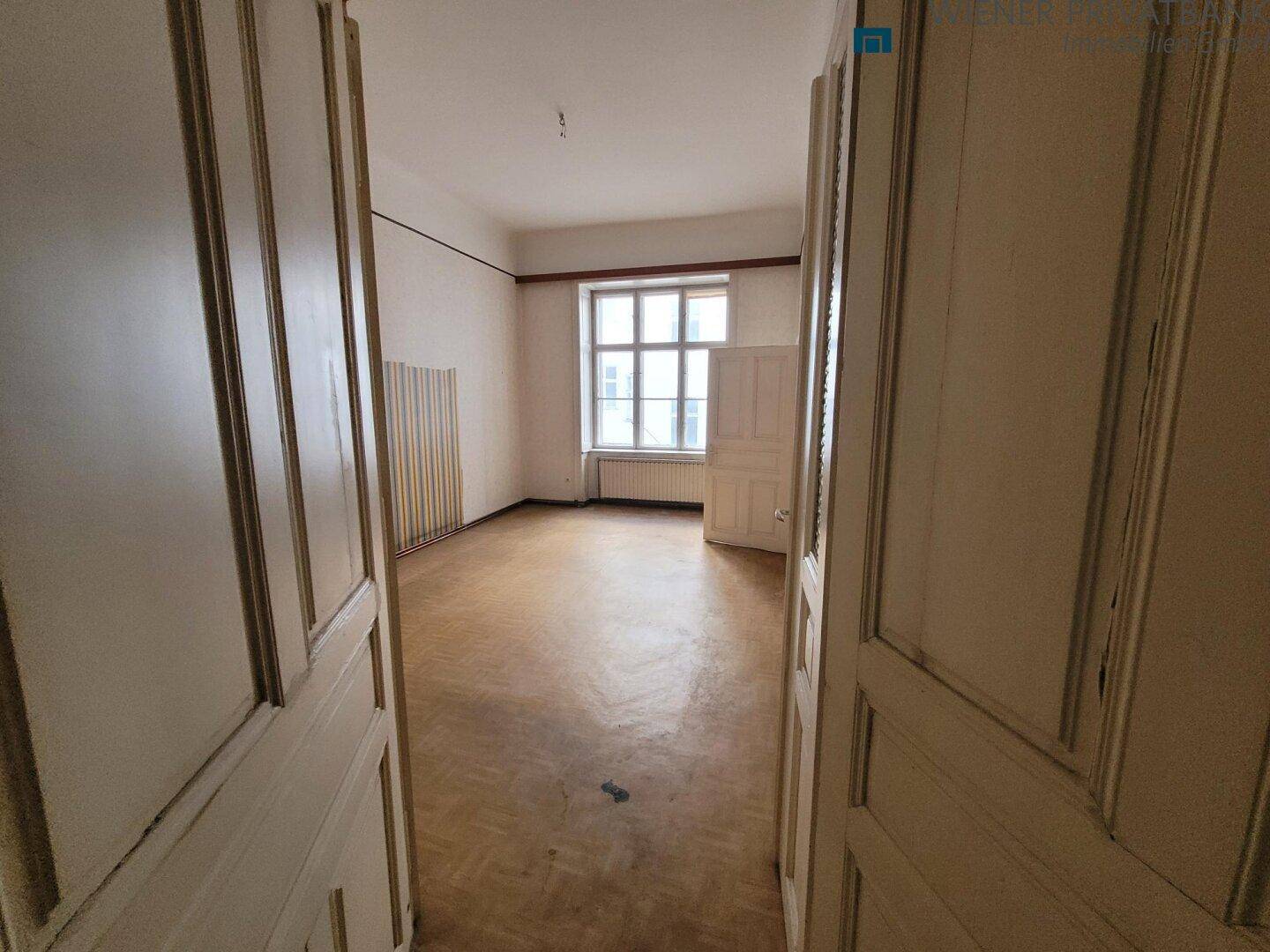Geräumiger Raum mit Fischgrätparkett und Fenster, gesehen durch eine offene Flügeltür, renovierungsbedürftig.