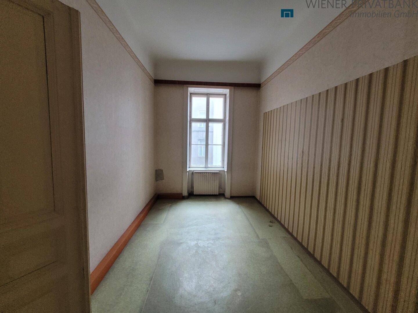 Schmaler Raum mit gestreifter Tapete, Fenster und altem Teppichboden, der eine Sanierung benötigt.