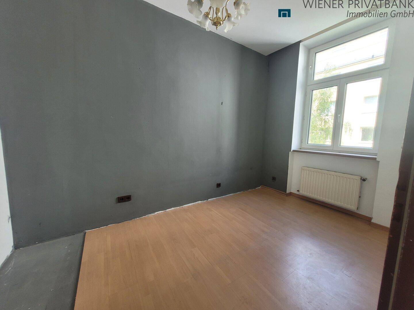 Unmöbliertes Zimmer mit Holzboden, grauer Akzentwand und Fenster.