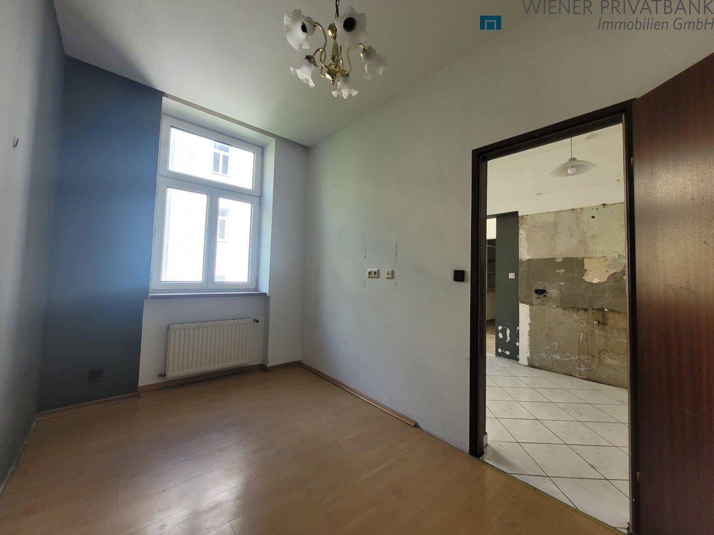 Leeres Zimmer mit Holzboden und Blick in einen renovierungsbedürftigen Bereich.