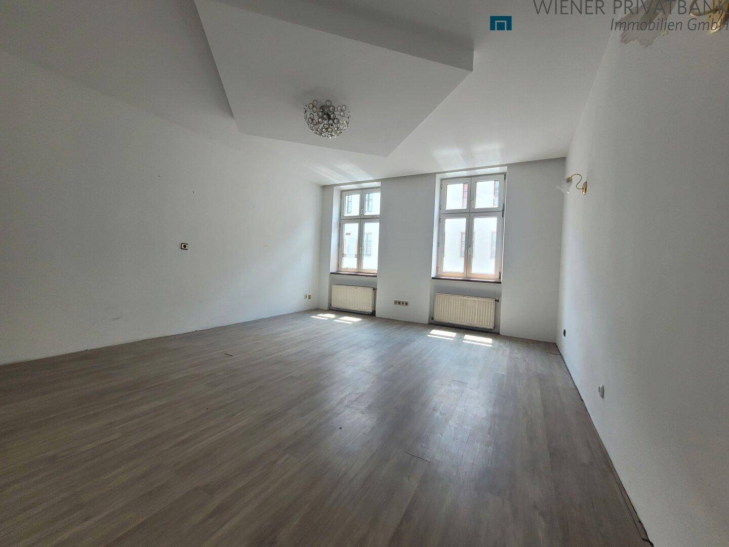 Geräumiges, unmöbliertes Zimmer mit hellem Laminatboden und zwei großen Fenstern.