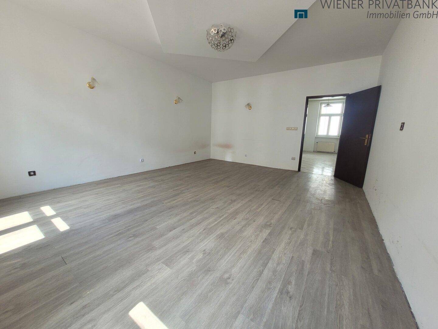 Großes, leeres Zimmer mit Laminatboden und viel Tageslicht.