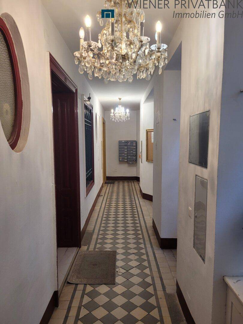 Langer Flur mit gemusterten Fliesen, zwei Kronleuchtern und Briefkästen an der Wand.