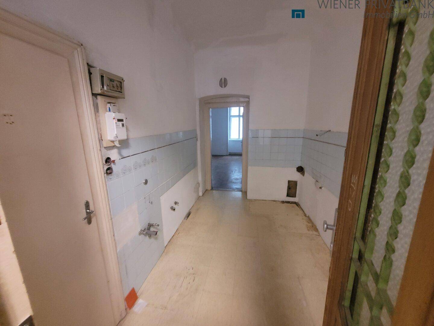 Leere Küche mit alten Fliesen und sichtbaren Anschlüssen, bereit für eine Renovierung.