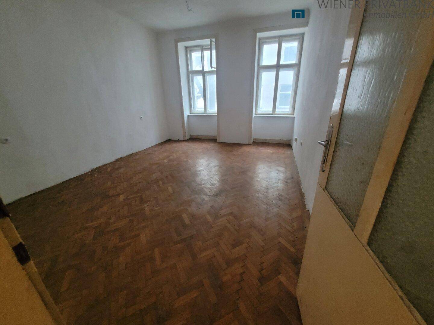 Leeres Zimmer mit Fischgrätparkett und zwei hohen Fenstern, bereit zur Gestaltung.