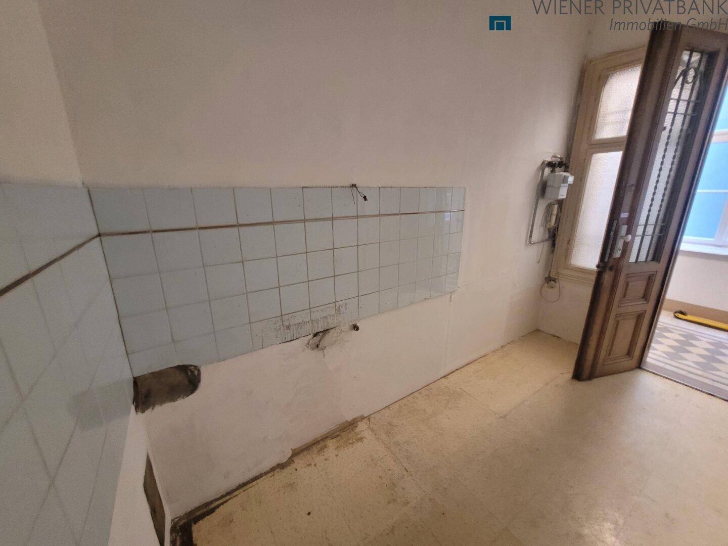 Leere Küche mit alten Fliesen und sichtbaren Anschlüssen, bereit für eine Renovierung.
