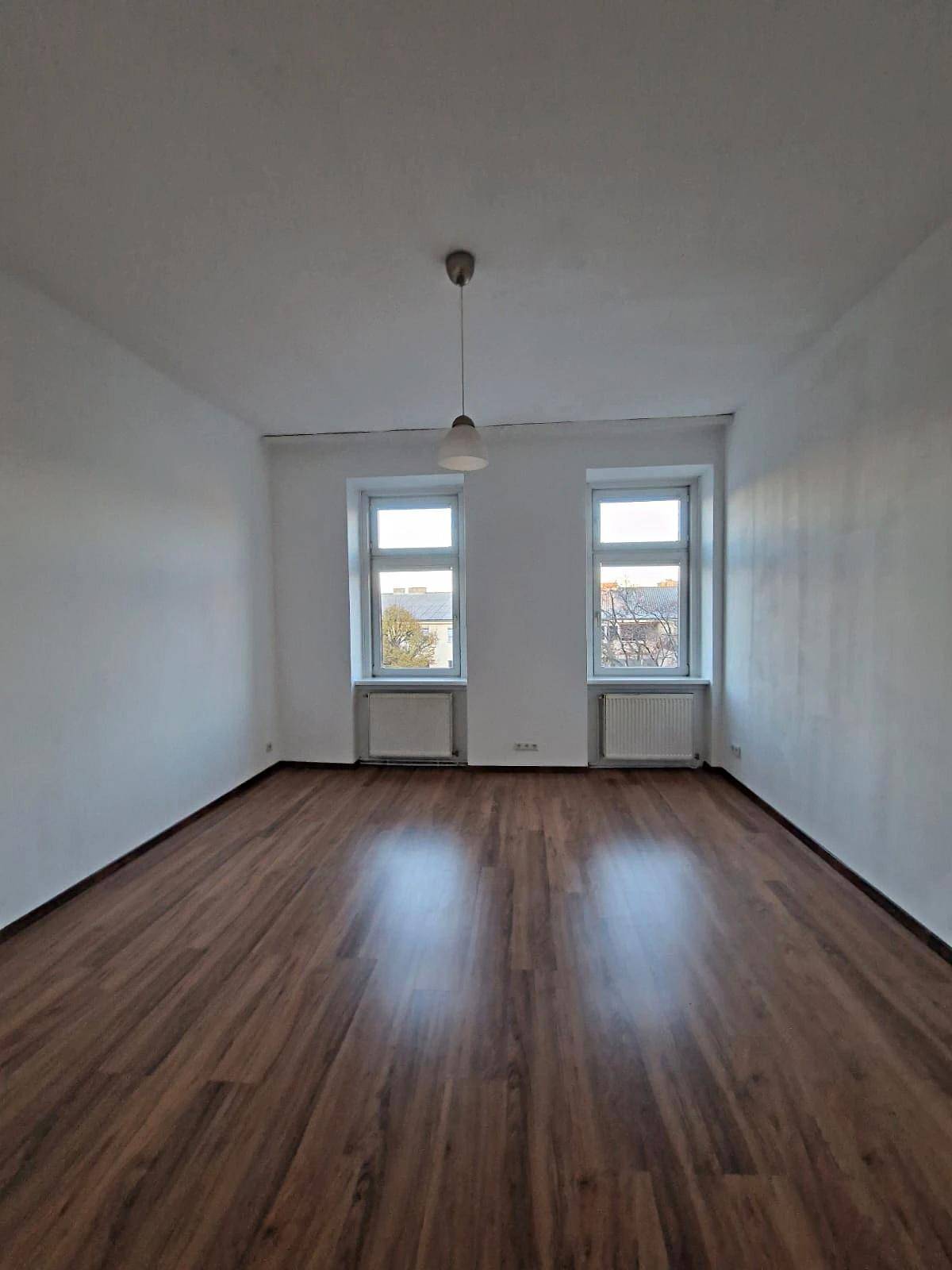 Geräumiges Zimmer mit zwei Fenstern und glänzendem Holzparkett, bietet viel Tageslicht.
