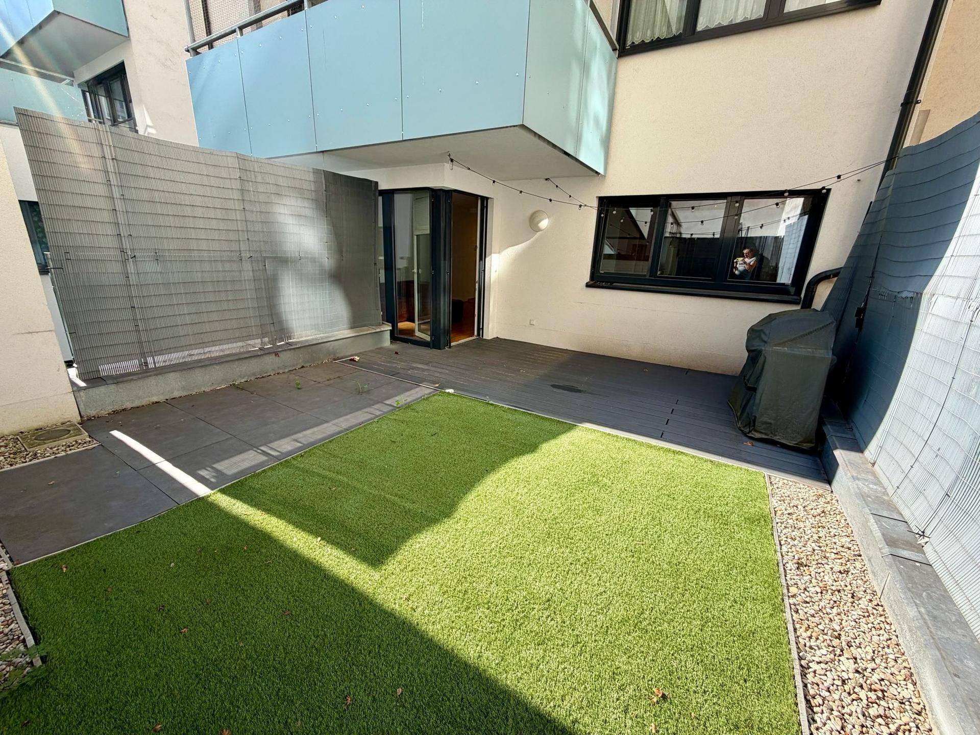 Sonniger Außenbereich mit Kunstrasen, gepflasterter Terrasse und Grillplatz.