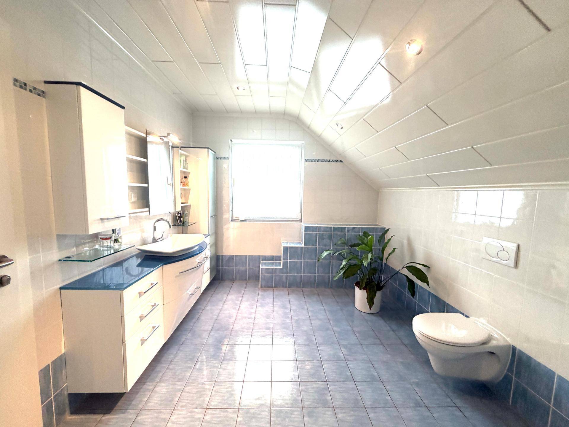 Geräumiges Badezimmer im Dachgeschoss mit blauen Fliesen, Fenster und wandhängendem WC.