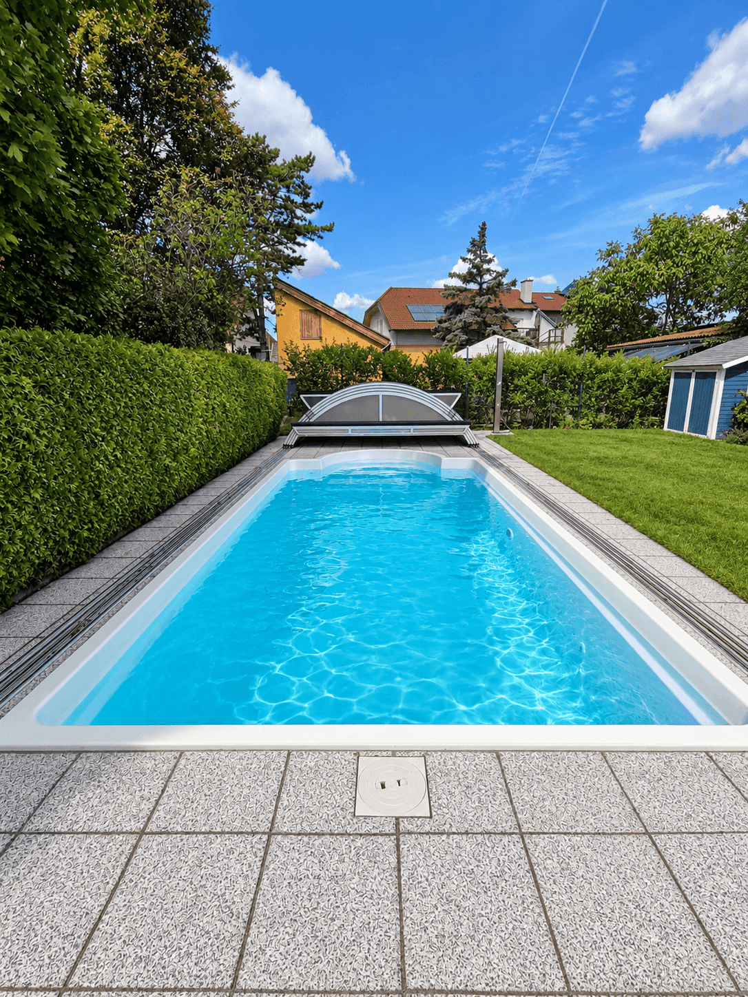 Langer, rechteckiger Swimmingpool im Garten mit gepflasterter Umrandung und grüner Hecke.