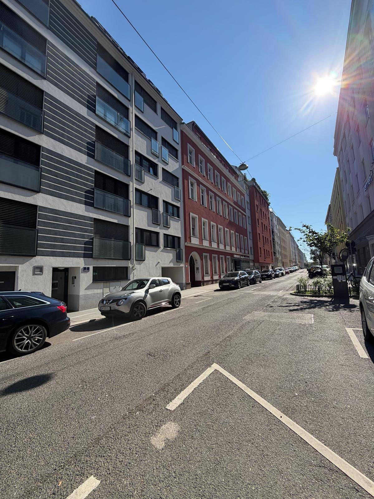 Sonnige Stadtstraße mit geparkten Autos und verschiedenen Wohngebäuden unter blauem Himmel.