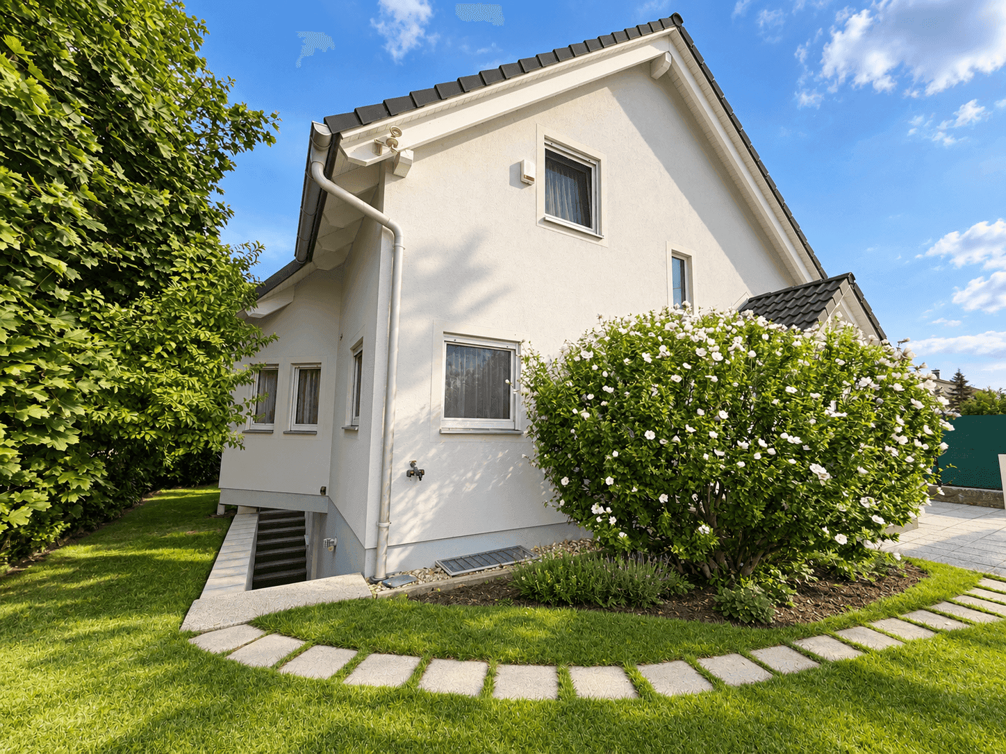 Seitenansicht eines weißen Einfamilienhauses mit gepflegtem Garten und Kellerzugang.