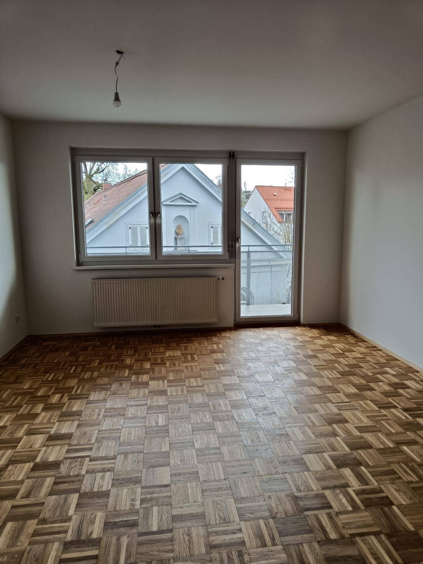 Heller, leerer Raum mit Parkettboden, großem Fenster und Zugang zu einem Balkon.