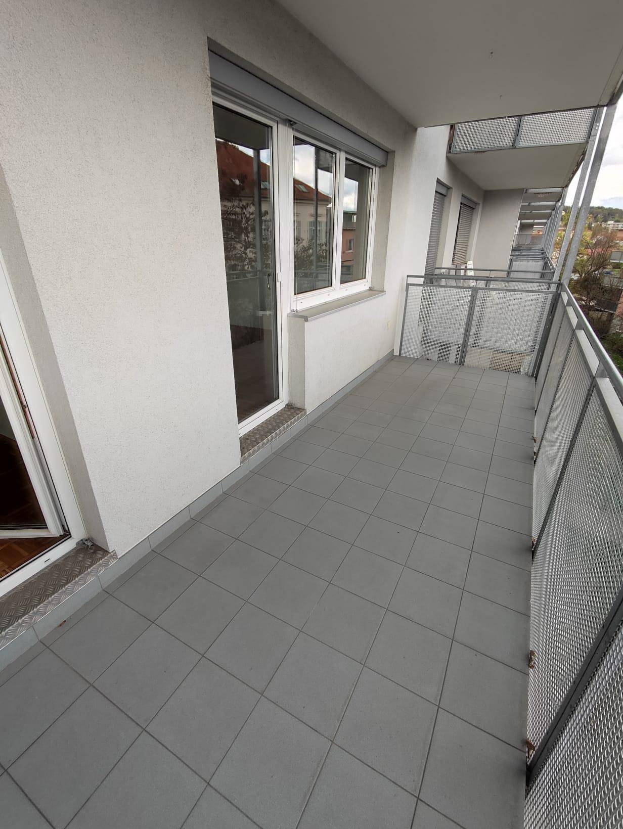 Langer Balkon mit grauen Fliesen, robustem Geländer und Rollläden an den Fenstern.