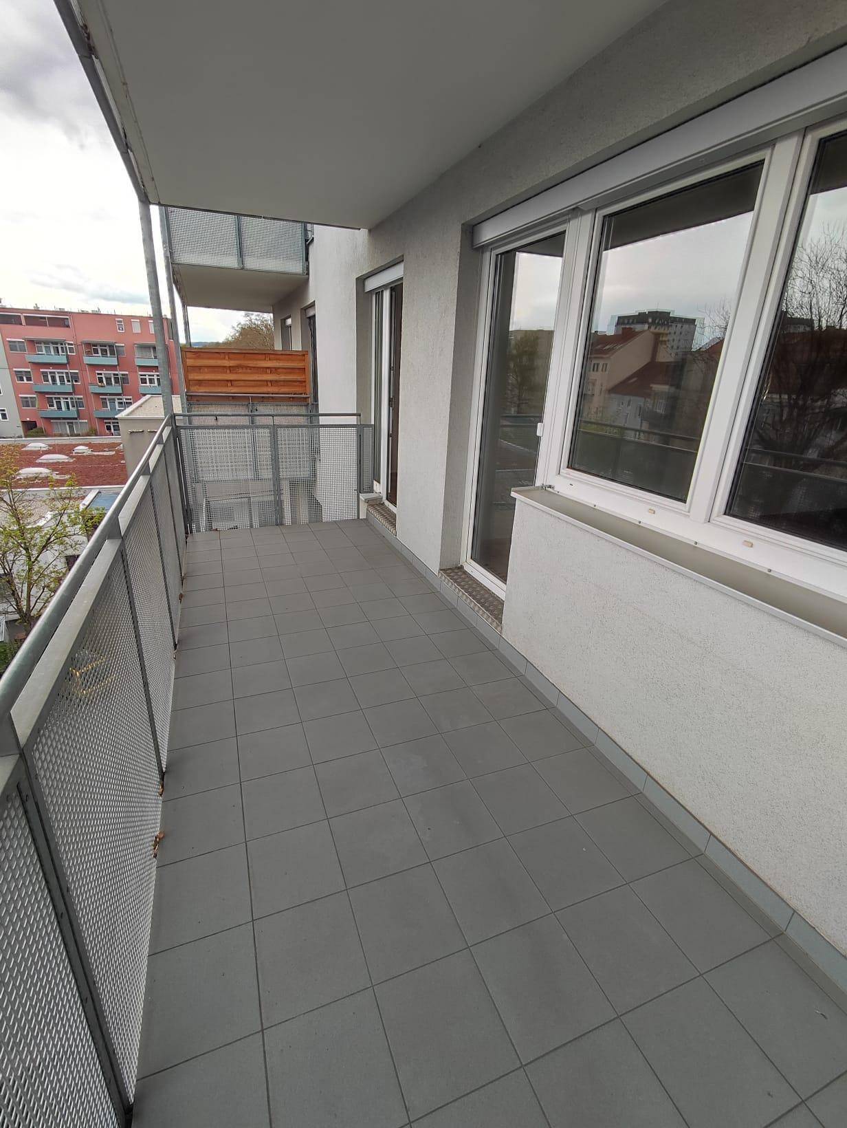 Geräumiger Balkon mit grauen Fliesen, Metallgeländer und Blick auf die umliegenden Gebäude.