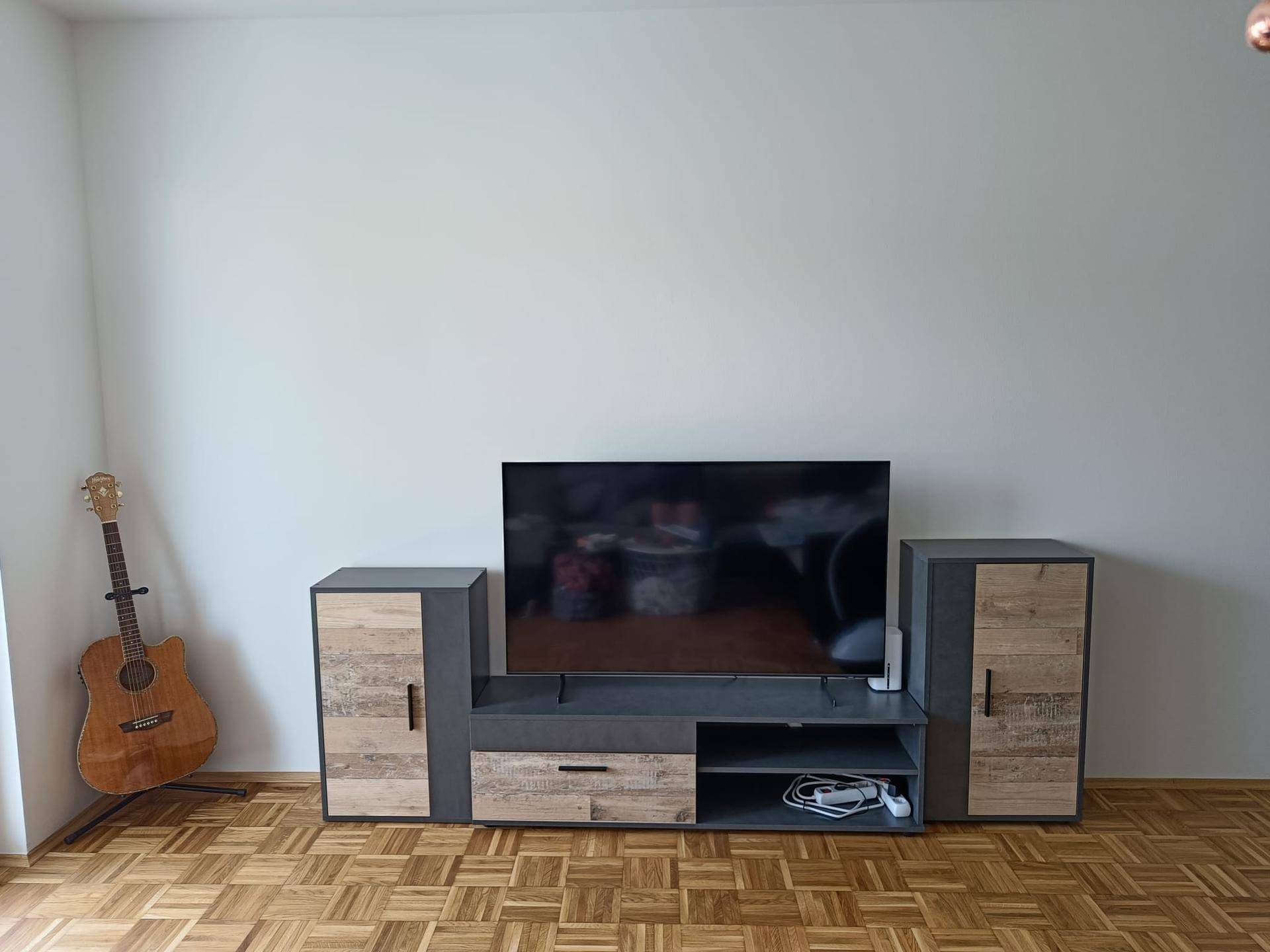 Wohnzimmer mit TV-Möbeln, Fernseher und Gitarre an einer schlichten weißen Wand.