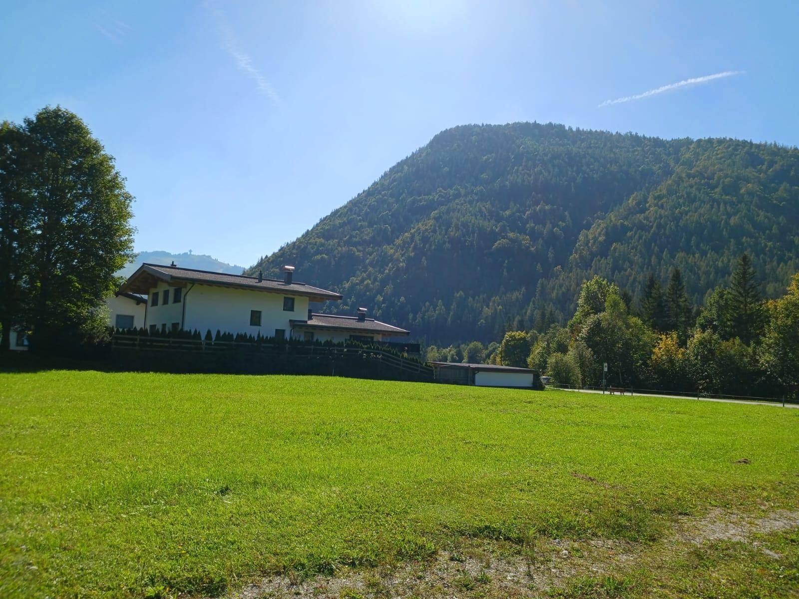 Idyllische Szene mit einem Haus am Rande einer grünen Wiese und einem bewaldeten Berg im Hintergrund.