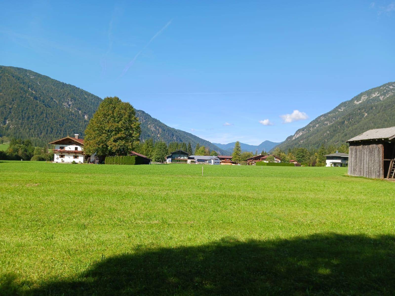 Weitläufige grüne Landschaft mit Häusern in einem Tal, umgeben von bewaldeten Bergen unter blauem Himmel.