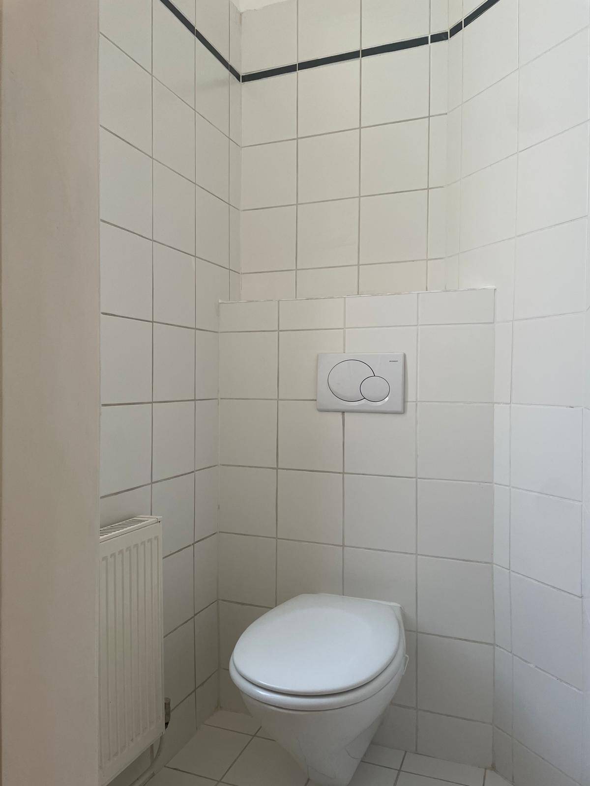 Separates WC mit wandhängender Toilette und weißen Fliesen mit schwarzem Akzentstreifen.
