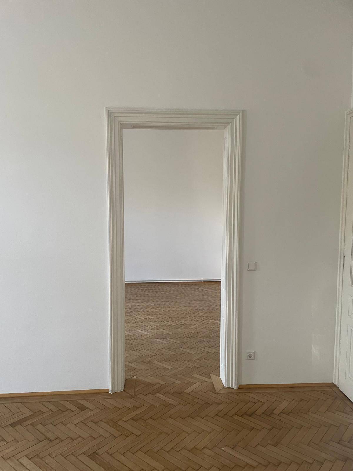 Leerer Raum mit Fischgrätparkett und einer offenen Tür, die den Blick in einen weiteren Raum freigibt.
