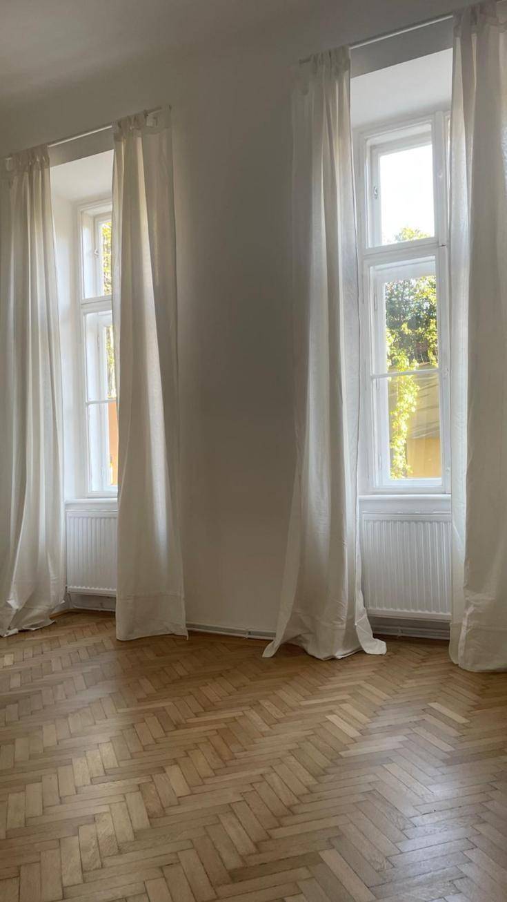 Lichtdurchfluteter Raum mit zwei Fenstern, weißen Vorhängen und elegantem Fischgrätparkett.