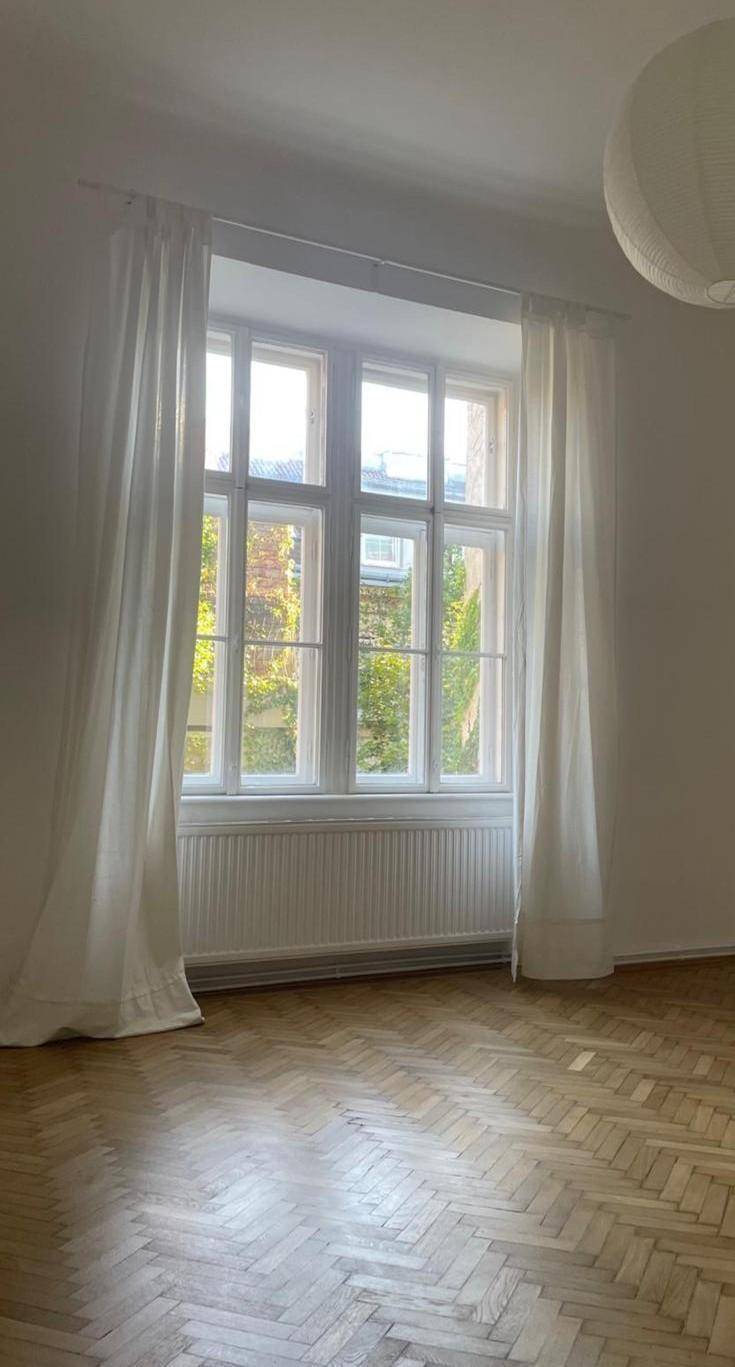Heller Raum mit großem Fenster, weißen Vorhängen und klassischem Fischgrätparkettboden.