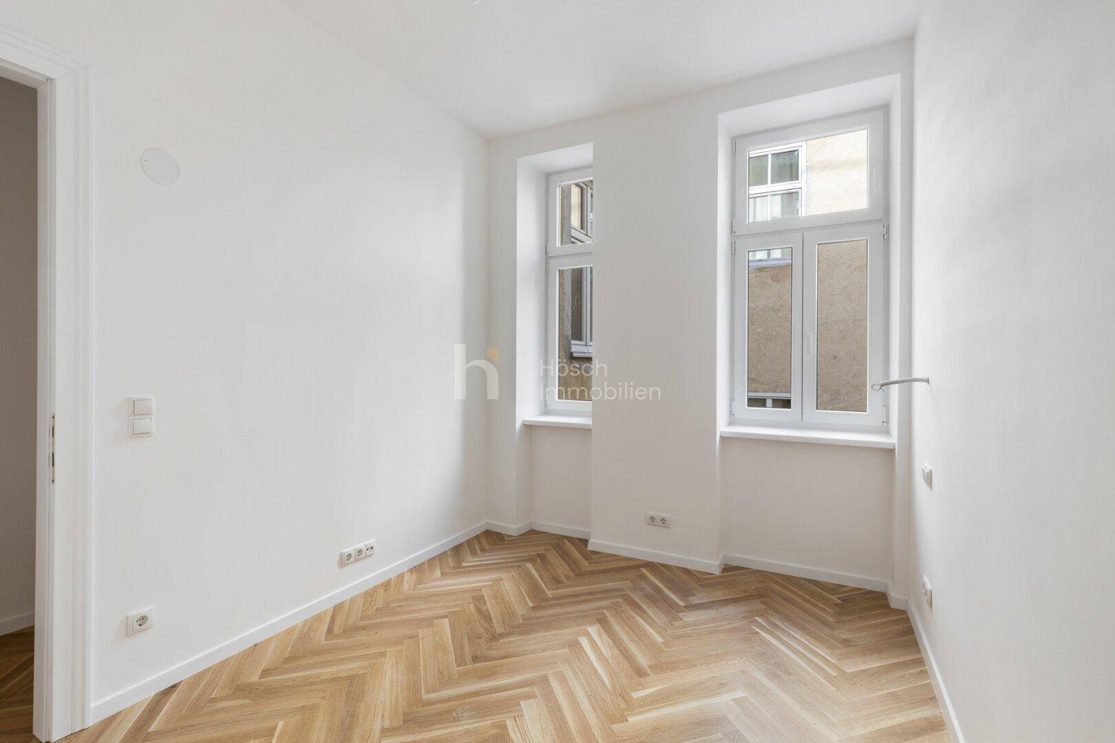 Heller Raum mit Fischgrätparkett und zwei hohen Fenstern, ideal für ein Schlaf- oder Arbeitszimmer.