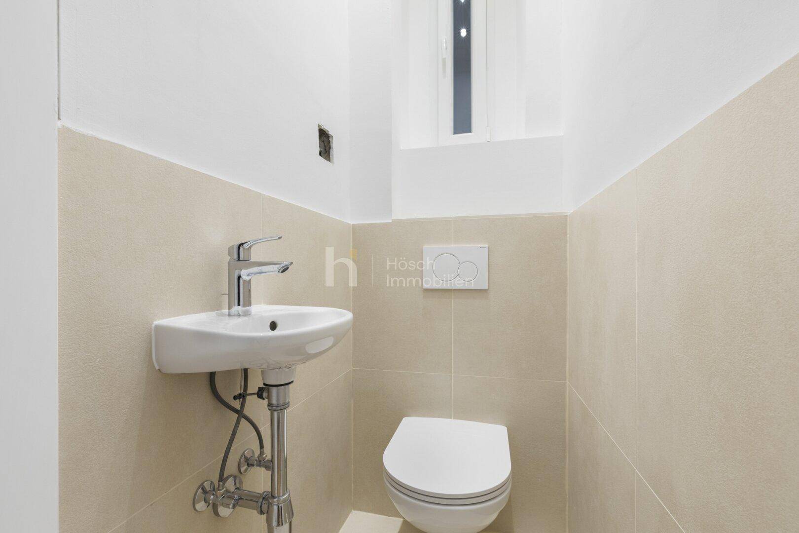 Separates Gäste-WC mit modernem Wand-WC, kleinem Waschbecken und beigefarbenen Fliesen.