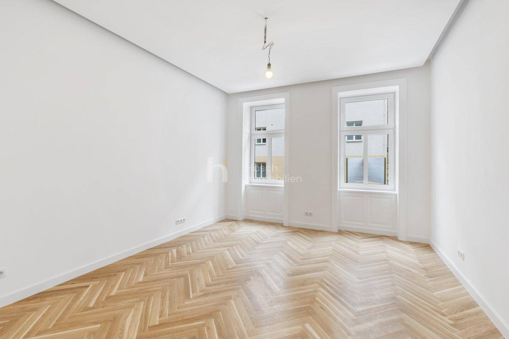 Geräumiger, leerer Raum mit Fischgrätparkett und viel Tageslicht durch hohe Fenster.