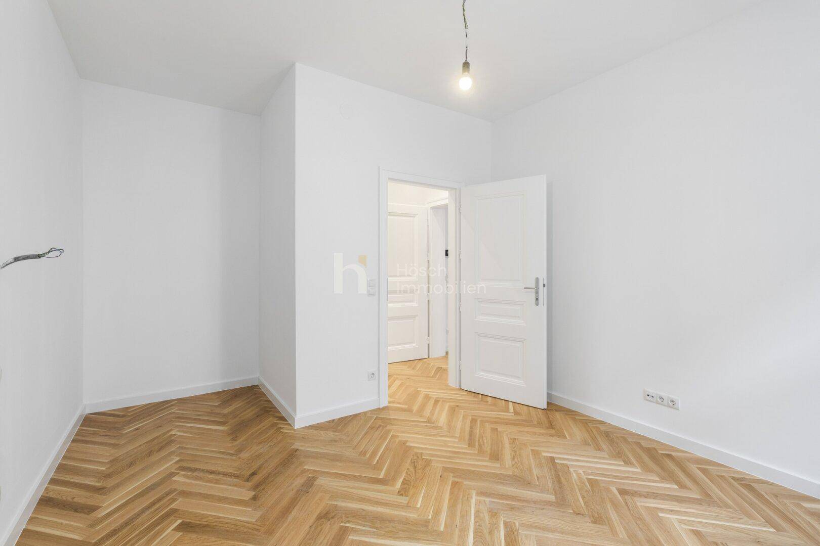 Heller, unmöblierter Raum mit Fischgrätparkett und offener Tür zu einem weiteren Zimmer.