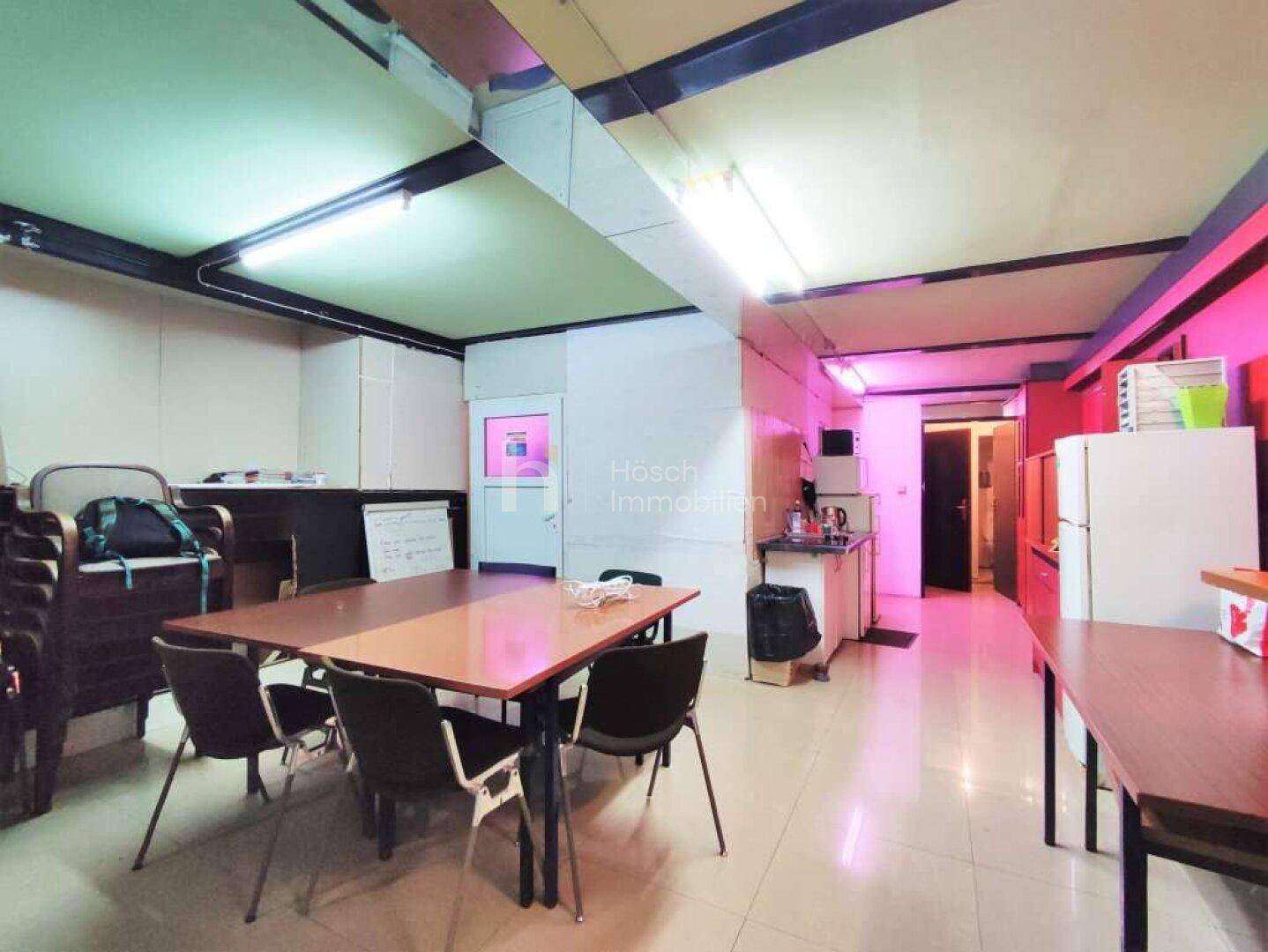 Mehrzweckraum mit Tischen, Stühlen und einer kleinen Küchenzeile, beleuchtet mit Neonlicht.