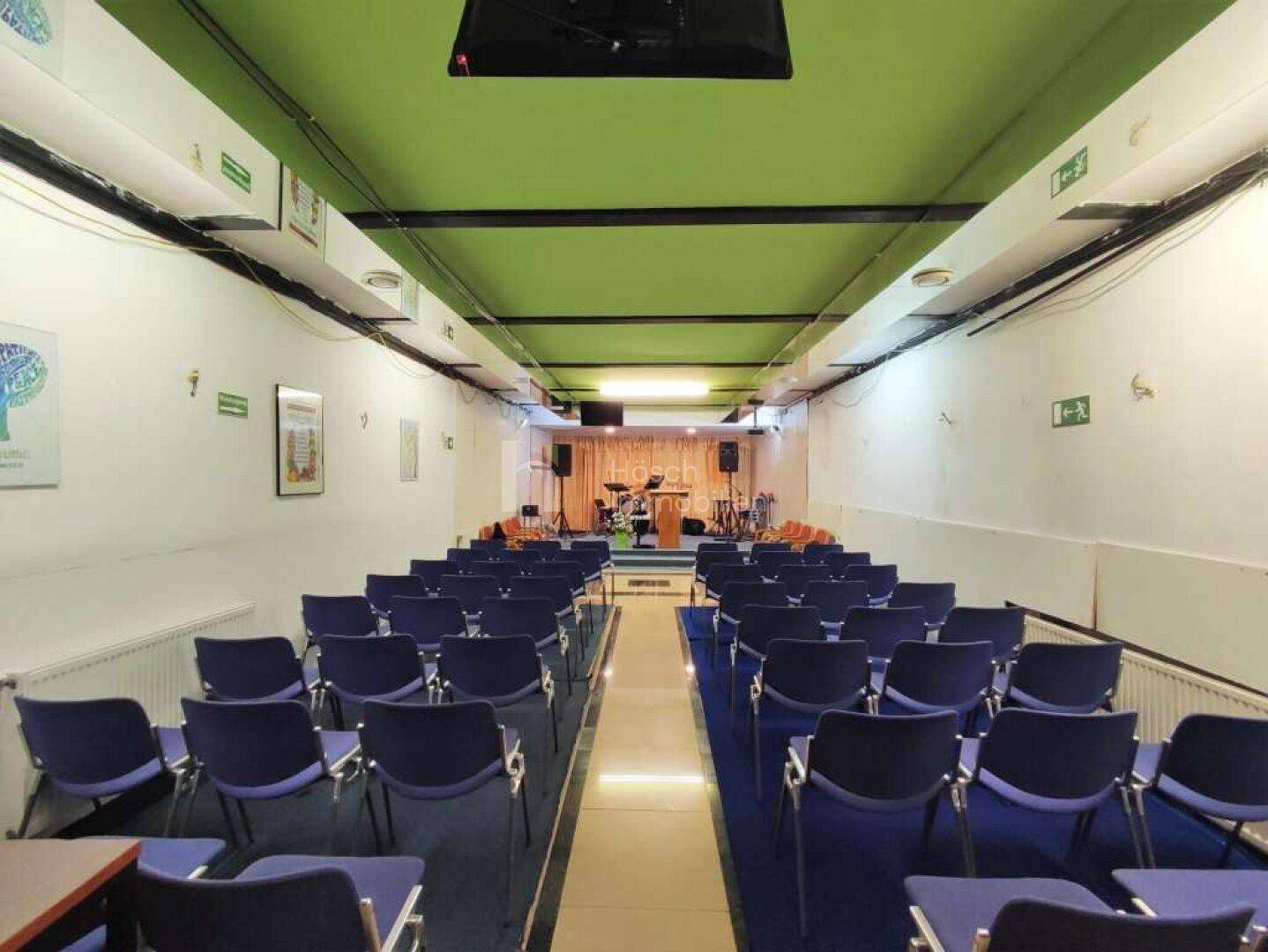 Großer Versammlungsraum mit blauen Stühlen, Bühne und auffälliger grüner Decke.