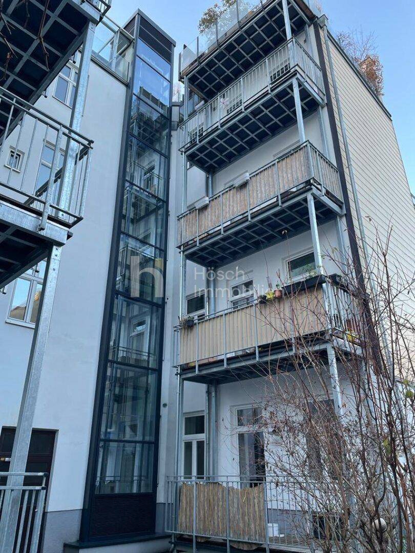 Rückseite des Gebäudes mit mehreren Balkonen und einem außenliegenden Glasaufzug für modernen Komfort.