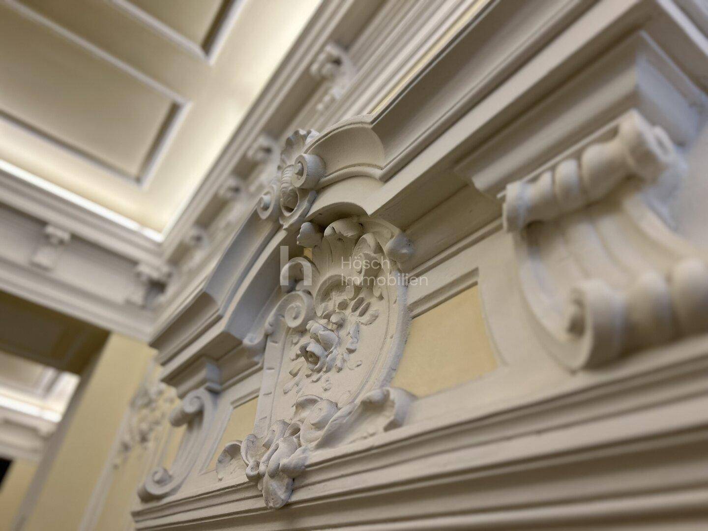 Nahaufnahme einer eleganten Stuckverzierung an Decke und Wand, die den aristokratischen Stil des Gebäudes unterstreicht.