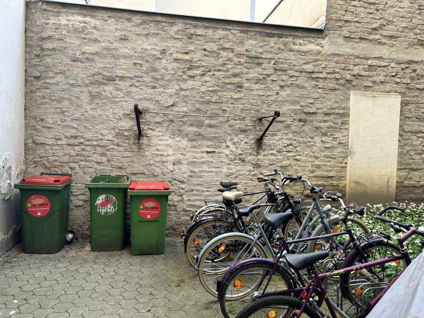 Gepflasterter Innenhofbereich mit Fahrradabstellplätzen und Mülltonnen vor einer Ziegelwand.
