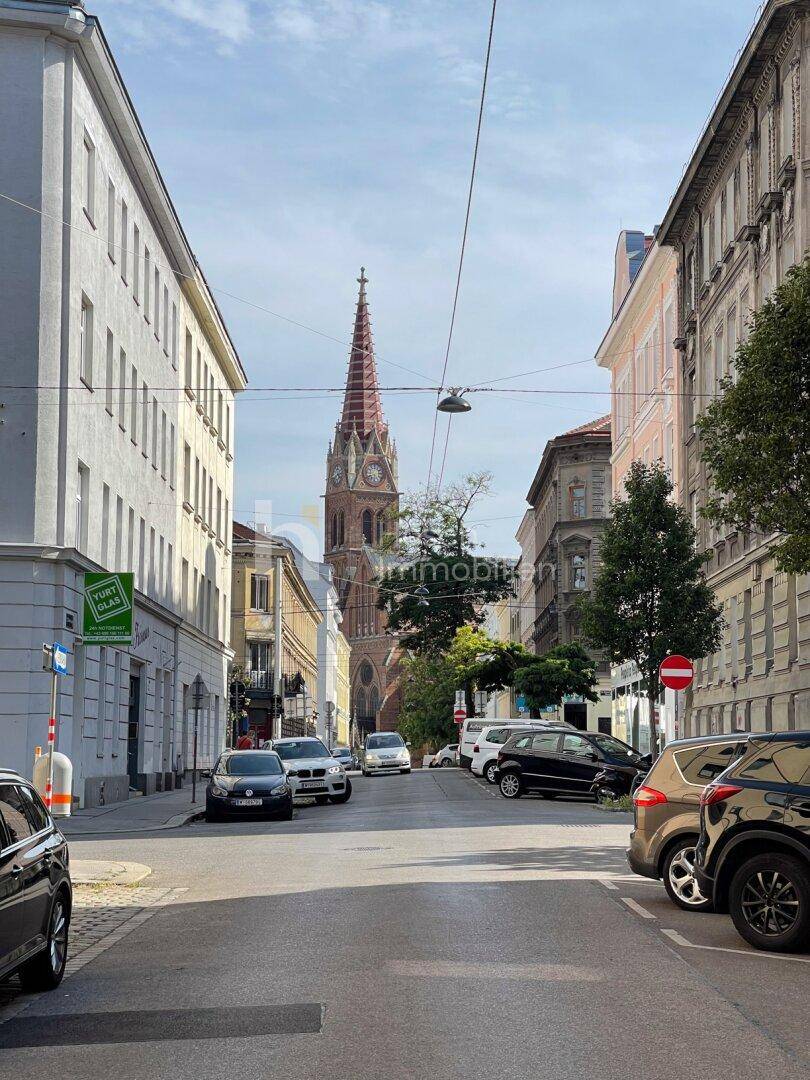 Blick entlang einer Stadtstraße auf einen markanten Kirchturm, umgeben von Gebäuden und Autos.
