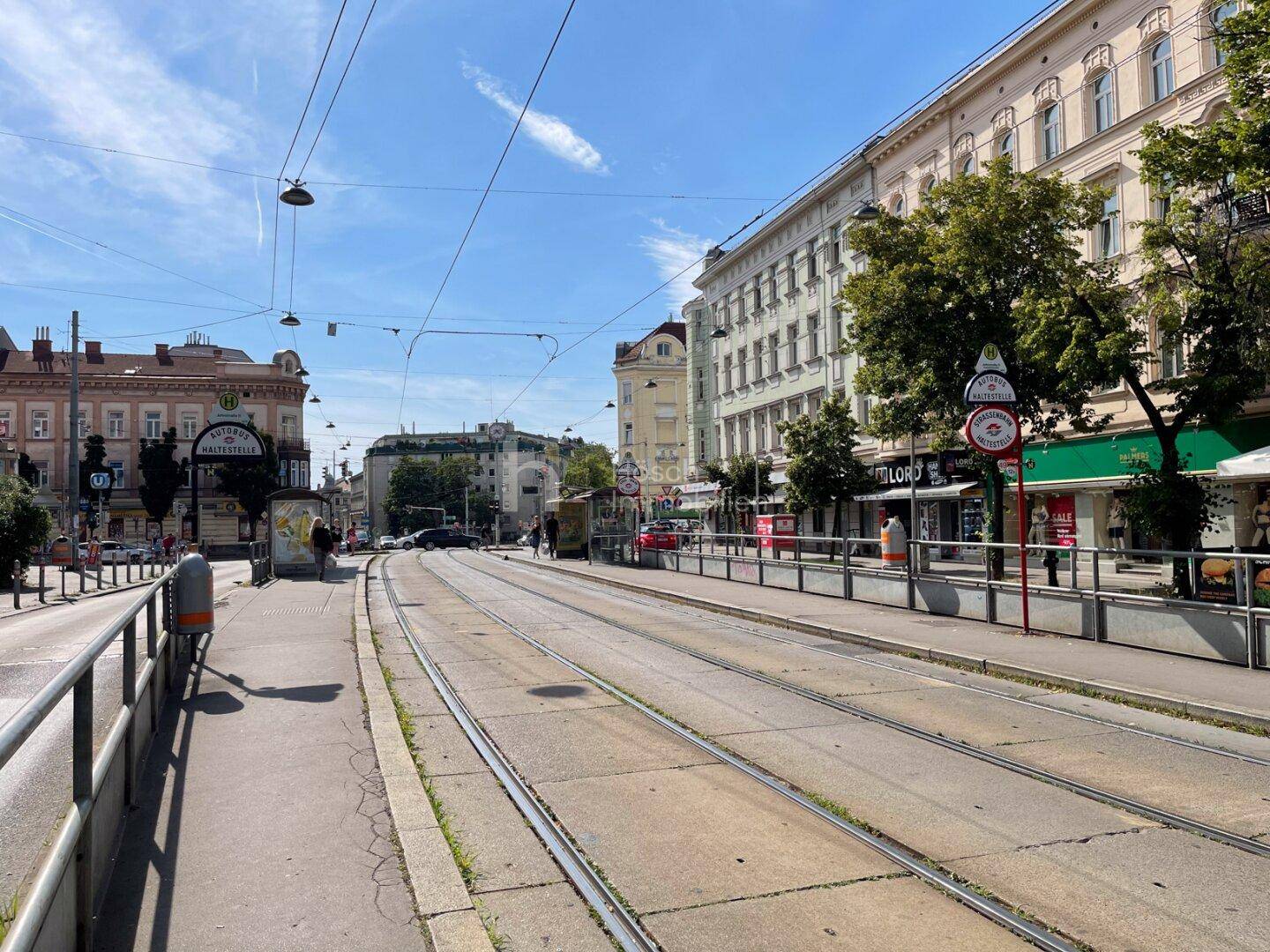 Weitläufige Stadtansicht mit Straßenbahnschienen, historischen Gebäuden und blauem Himmel.