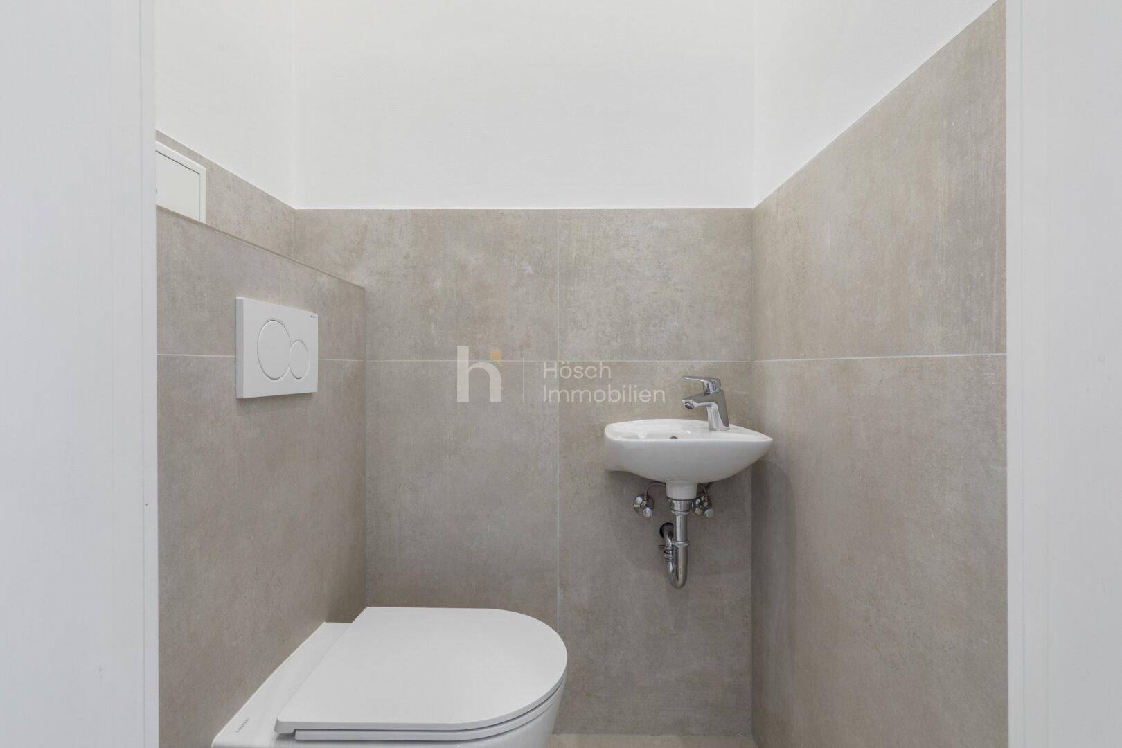 Gäste-WC mit Wand-WC, kleinem Waschbecken und modernen grauen Wandfliesen.