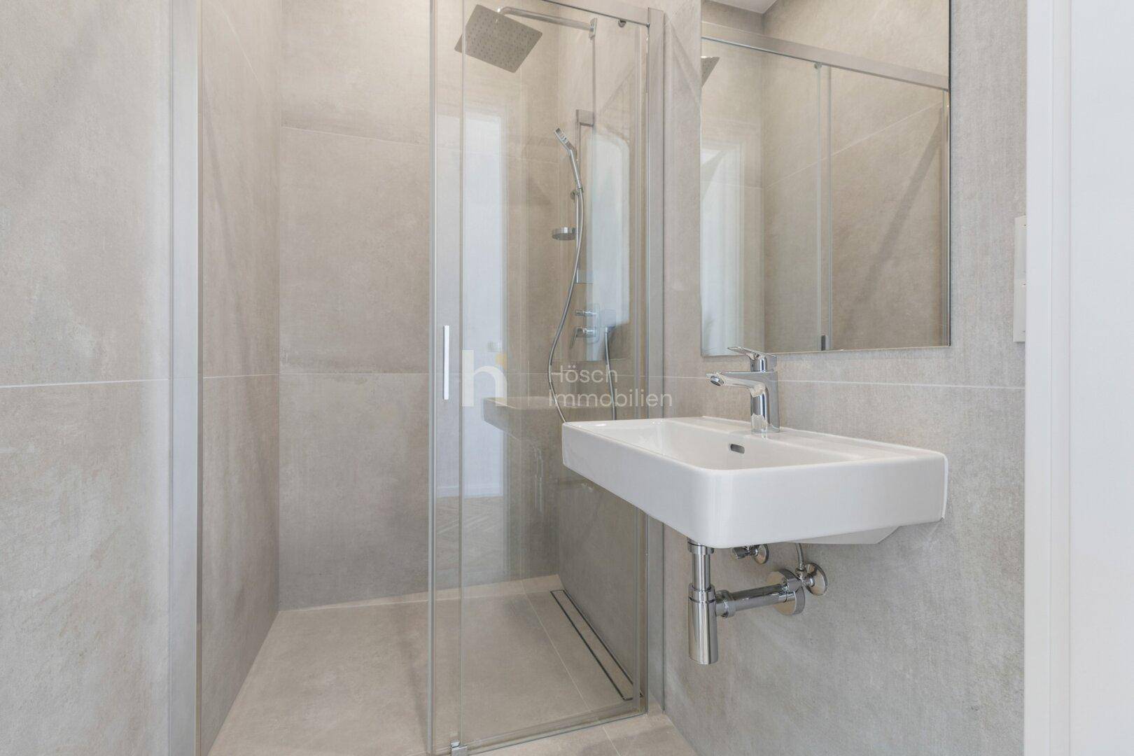 Modernes Badezimmer mit begehbarer Glasdusche, Wandwaschbecken und großen grauen Fliesen.
