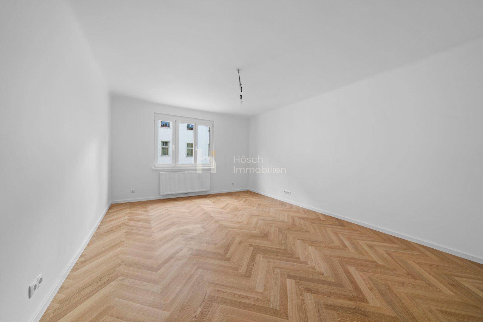 Leeres Zimmer mit Fischgrätparkett und Fenster, das viel natürliches Licht hereinlässt.
