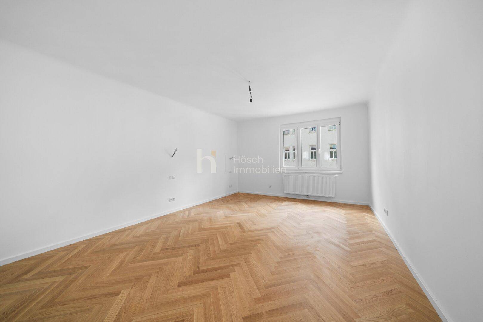 Helles, leeres Zimmer mit Fischgrätparkett und Fenster, das eine angenehme Atmosphäre schafft.